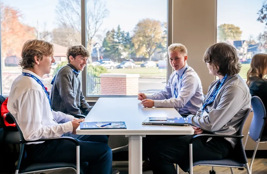 Four students talk at a table next to a window inside.